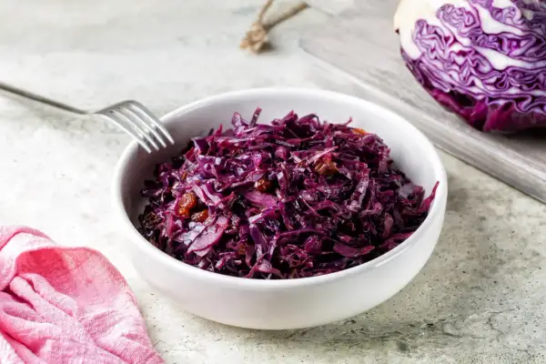 Un bol de chou rouge recette, garni de raisins secs, prêt à être dégusté. Une moitié de chou rouge est visible sur une planche à découper en arrière-plan.