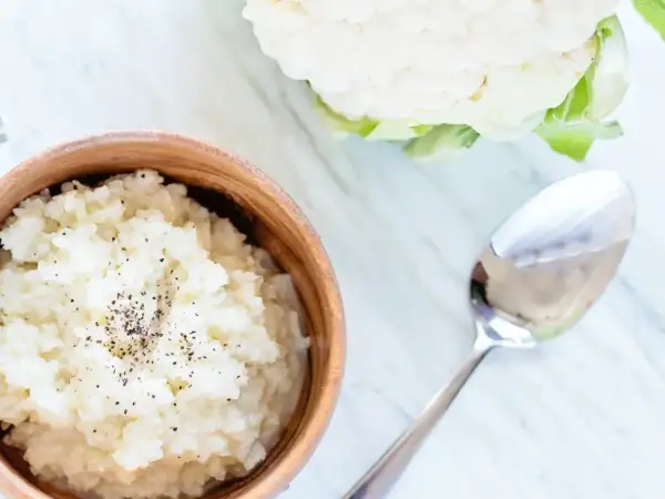 Pyszne puree z kalafiora w drewnianej misce, posypane pieprzem. Obok leży łyżka, a w tle świeży kalafior.