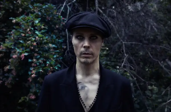 Ville Valo w czarnym kaszkiecie i marynarce, z intensywnym makijażem oczu, stoi na tle ciemnej, leśnej roślinności.
