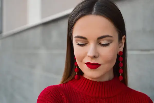 Une femme arbore un beau maquillage Noël, avec des lèvres rouge vif et des yeux dorés, portant un pull rouge et des boucles d'oreilles assorties.