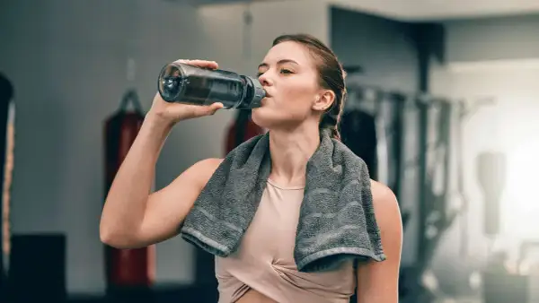 Eine Frau trinkt nach dem Training aus ihrer Trinkflasche. Sie hat ein Handtuch um den Hals.