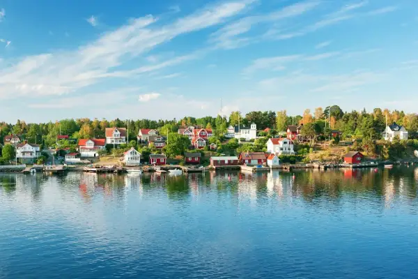 Vackra röda och vita hus längs en grönskande kustlinje, speglande sig i det blå vattnet. En idyllisk vy av sommar i Stockholm.