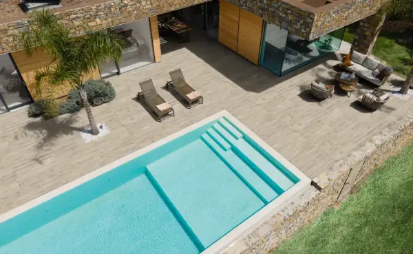 Una piscina azzurra e una moderna pavimentazione esterna in legno creano un'oasi di relax con arredi da esterno e una palma.