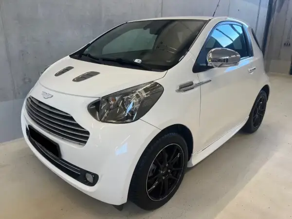 Biały Aston Martin Cygnet z czarnymi felgami i chromowanymi detalami, prezentujący się elegancko w garażu.