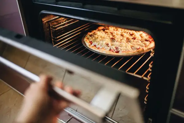Ręka otwiera piekarnik, w którym trwa pieczenie pizzy. Gorące wnętrze rozświetla apetyczną potrawę.