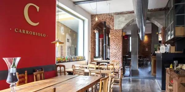 Interno di un locale con tavoli in legno e mattoni a vista. Sulla parete rossa, il logo "C CARROBIOLO" e una vetrina con tante bottiglie di birra.