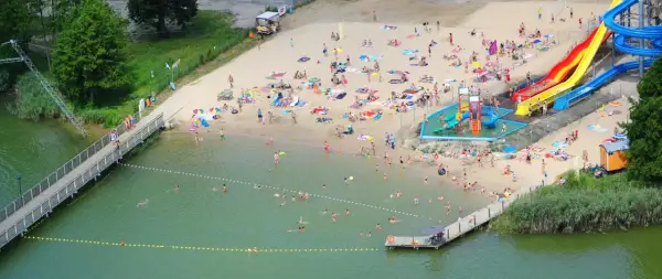 Wakacyjny dzień nad jeziorem margonińskim. Ludzie opalają się na plaży, kąpią w wodzie i korzystają ze zjeżdżalni wodnych.