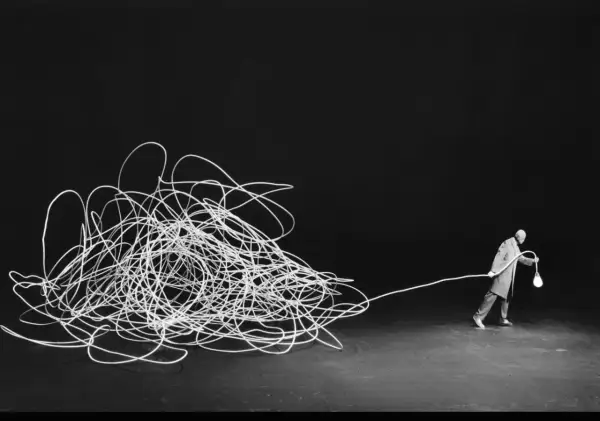 Człowiek z żarówką, jak w pracach Gilbert Garcin, ciągnie za sobą plątaninę białego sznurka na czarnym tle.