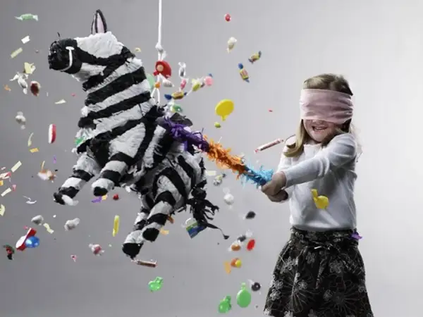 Zabawa w wypełnienie piniaty w kształcie zebry. Dziewczynka z zawiązanymi oczami próbuje ją rozbić, a cukierki i konfetti lecą w powietrzu.