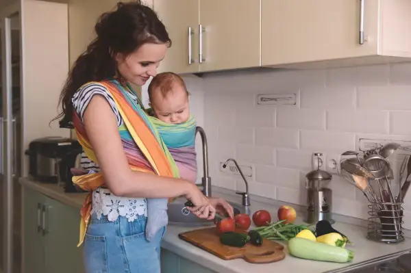 Mamma förbereder mat vid amning i köket, med en bebis i bärsjal. Grönsaker ligger på bänken.