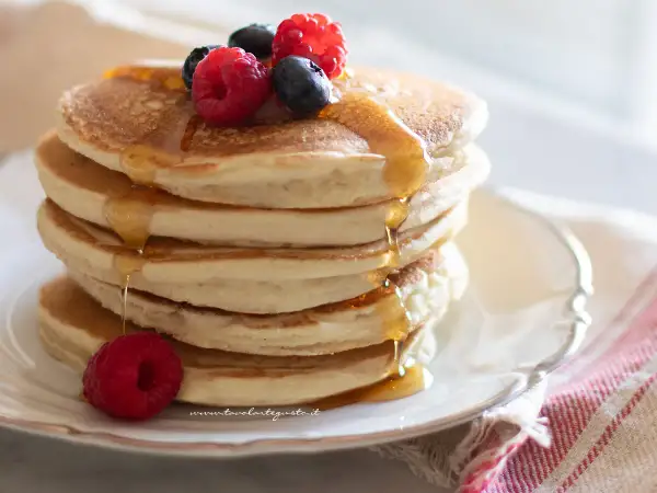 Pancakes senza uova dorati, impilati e inzuppati di sciroppo, guarniti con lamponi e mirtilli freschi.