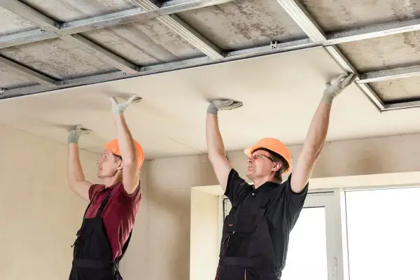 Deux ouvriers en casque orange installent des plaques de plafond, une étape clé de la rénovation plafond.