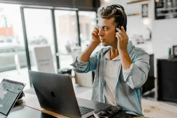 A young man, likely one of the new generation of music managers, adjusts his headphones while working on his laptop in a cafe.