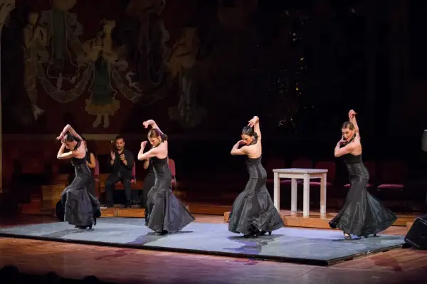 Cuatro bailarinas de flamenco, con vestidos negros voluminosos, ejecutan pasos apasionados. Un hombre aplaude al fondo.