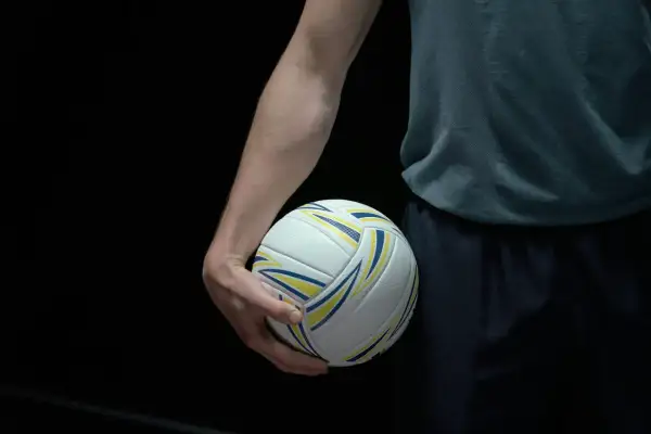 Dłoń zawodnika trzyma piłkę do siatkówki. Ciemne tło podkreśla biało-żółto-niebieską piłkę, gotową do gry w siatkówkę mężczyzn plusliga.