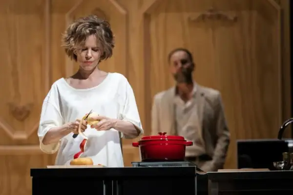 La locandiera recensioni: donna sbuccia una mela, pentola rossa sul fuoco, uomo sullo sfondo.