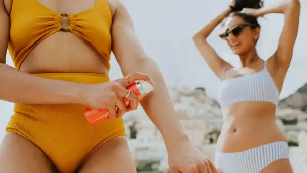 Sonnenschutz auftragen: Eine Frau sprüht sich mit Sonnencreme LSF 30 oder 50 ein, während ihre Freundin im Hintergrund die Sonne genießt.
