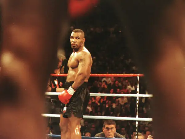 Mike Tyson full body, standing in a boxing ring, wearing red gloves and black shorts, with a crowd watching.