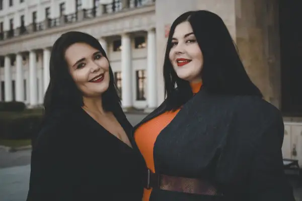 Dwie uśmiechnięte kobiety, uosobienie body positivity, pozują przed budynkiem z kolumnami.