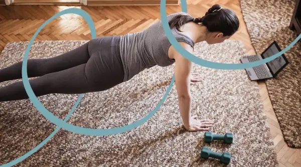 Kobieta w pozycji deski ćwiczy pilates w domu, patrząc na laptopa. Obok leżą hantle.