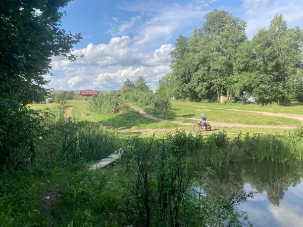 Osoba na motocyklu crossowym jedzie po szutrowej drodze wśród zieleni, obok stawu i drewnianego mostku.