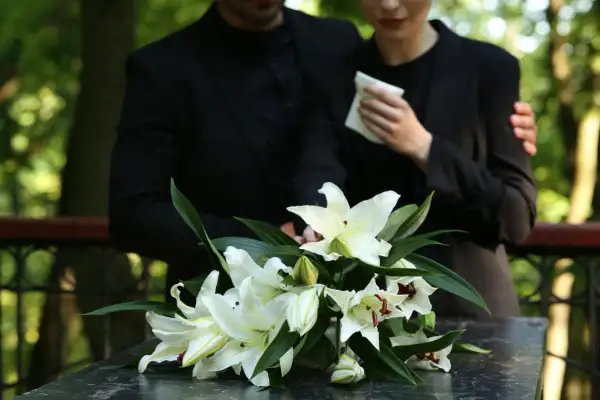 Białe lilie na ciemnym blacie, w tle para w żałobie.