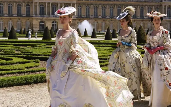 Kvinnor i magnifika **rococo fashion**-klänningar med blomstermönster promenerar i en formell trädgård framför ett palats.