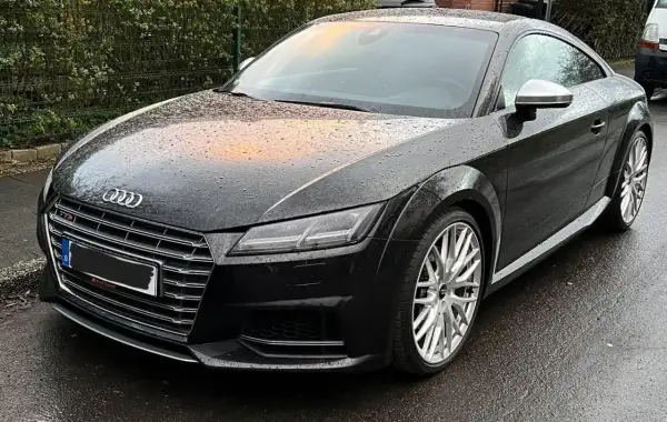 Negro Audi TT RS mojado por la lluvia, con reflejos naranjas en el capó. El coche está listo para la etapa 1 de su viaje.
