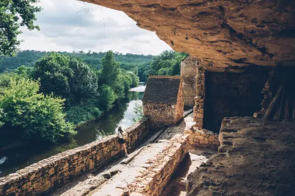 Une femme explore un village troglodytique en Dordogne, au bord d'une rivière paisible. Une belle idée de que faire en Dordogne.