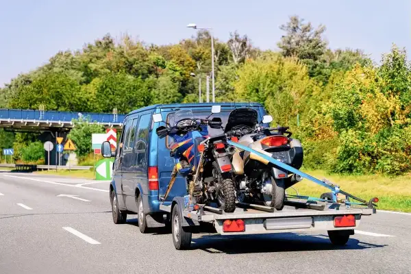 Jak przewieźć motocykl na zwykłej przyczepie? Dwa motocykle bezpiecznie zamocowane pasami na przyczepce za niebieskim vanem.