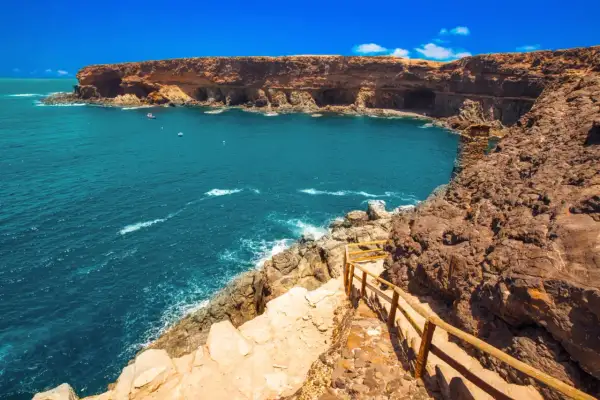 Fuerteventura na co uważać? Klify i turkusowe morze, ścieżka z drewnianą balustradą prowadzi w dół. Uważaj na nierówny teren.