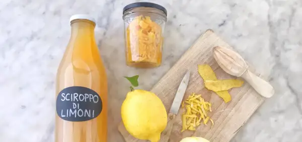 Bottiglia di sciroppo di limoni, limoni freschi, scorze e un coltello su tagliere. La sciroppo di limone ricetta della nonna è pronta!