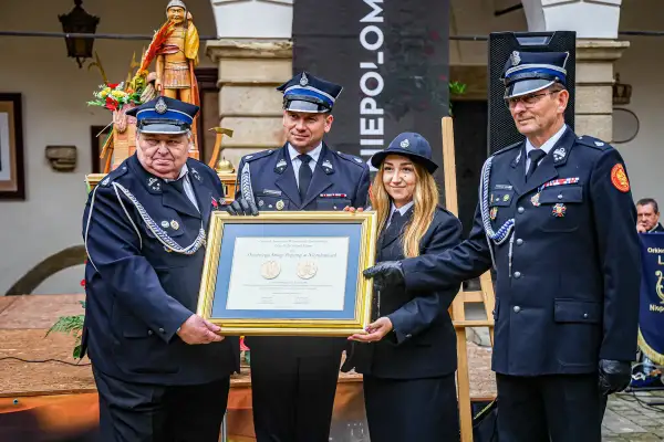 Uroczystość wręczenia nagród. Strażacy OSP w mundurach, z medalami, odbierają dyplom. Widać, jak nosić medale na mundurze OSP.