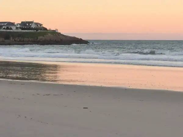 Ocaso en la playa areoura. Olas rompen suavemente en la arena dorada, reflejando el cielo rosado. Casas blancas coronan el acantilado.