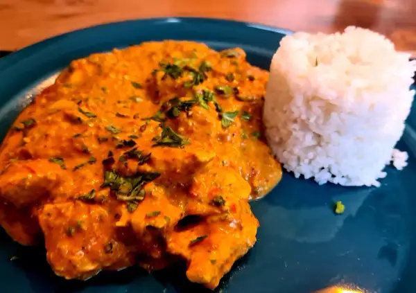 Ein Teller mit cremigem Hähnchen Tikka Masala, garniert mit frischer Petersilie, daneben ein Berg aus duftendem Reis.