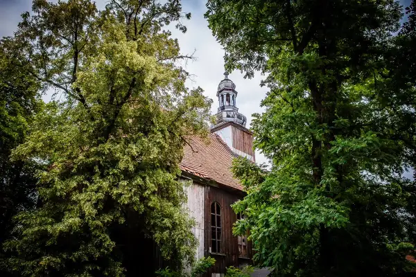 Stary kościół w Miejskiej Górce, otoczony bujną zielenią drzew, z charakterystyczną wieżą i drewnianą elewacją.