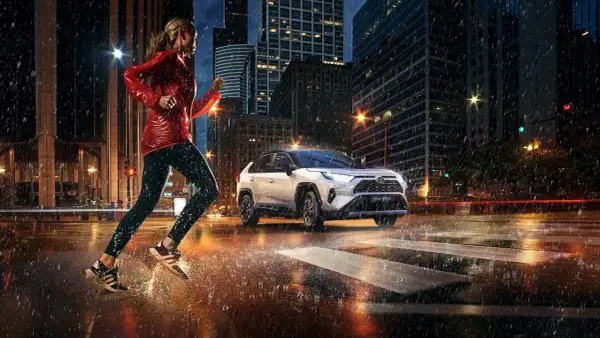 Kobieta biegnie w deszczu po mieście, obok niej jedzie biała Toyota RAV4 Hybrid. Zastanawiasz się, ile pali Toyota RAV4?