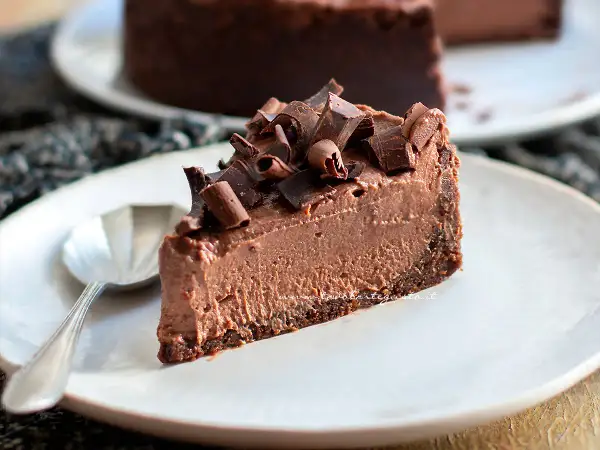 Una fetta di goloso cheesecake al cioccolato, guarnita con riccioli di cioccolato fondente, su un piatto bianco con un cucchiaino.