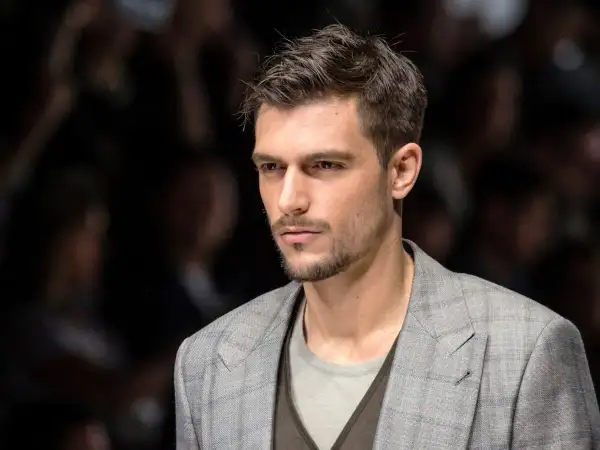Uomo con capelli spettinati corti, sguardo intenso, indossa una giacca a quadri grigia.
