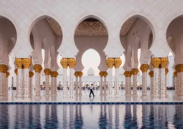 Un hombre camina bajo arcos blancos y dorados en Abu Dabi, reflejándose en el agua. Un visado Abu Dabi es el primer paso para disfrutar de esta maravilla.