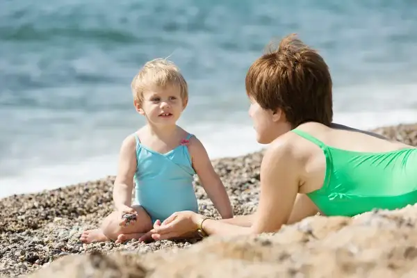 Mama i dziecko cieszą się wspólnym czasem nad morzem z dziećmi. Dziewczynka w turkusowym kostiumie bawi się kamykami, a mama w zielonym kostiumie leży obok.