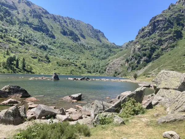 Lac de montagne entouré de montagnes verdoyantes et de rochers, sous un ciel bleu clair.