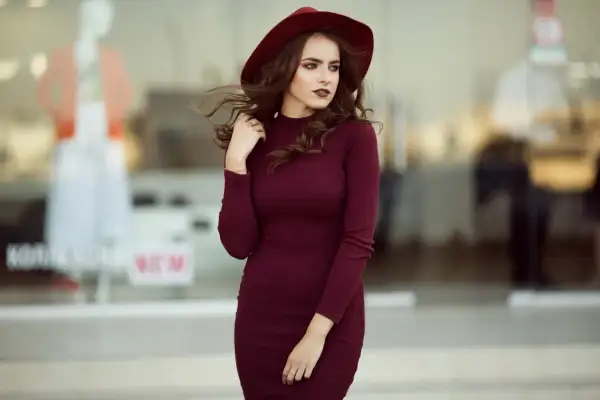 Elegancka bordowa sukienka i kapelusz tworzą stylową stylizację. Kobieta pozuje na tle witryny sklepowej.