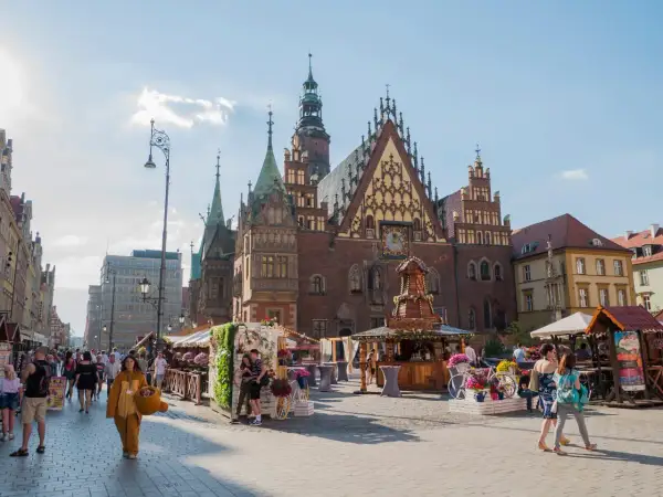 Tętniący życiem wrocławski Rynek, z Ratuszem w tle. Ludzie spacerują wśród kramów, a ktoś w stroju misia dodaje uroku temu miejscu.