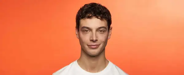 Hombre joven con cabello castaño rizado, luciendo una permanente ondulada. Sonríe levemente ante un fondo naranja vibrante.