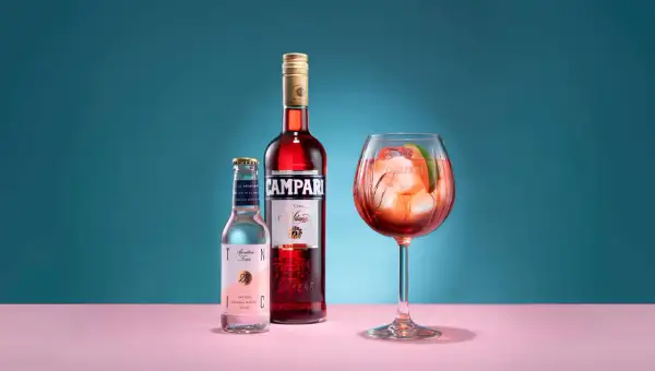 En Campari Spritz med is och lime i ett stort glas. Bredvid står en flaska Campari och en flaska tonic.