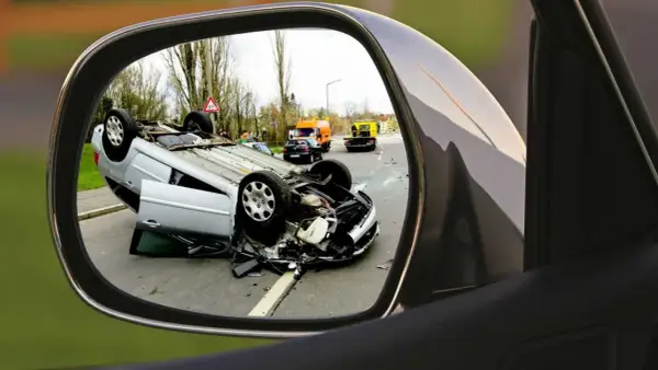 Auto w leasingu leży na dachu po wypadku. Widok w lusterku bocznym pokazuje szkody z OC sprawcy i pomoc drogową na drodze.