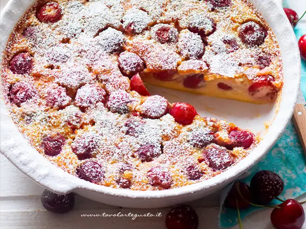 Clafoutis di ciliegie appena sfornato, cosparso di zucchero a velo, con una fetta mancante che rivela la morbida consistenza.