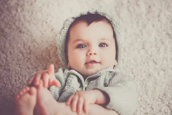 Bebé con ojos grandes y azules, vistiendo un gorro y suéter tejidos. Podría llamarse Gabriel, un nombre con g.