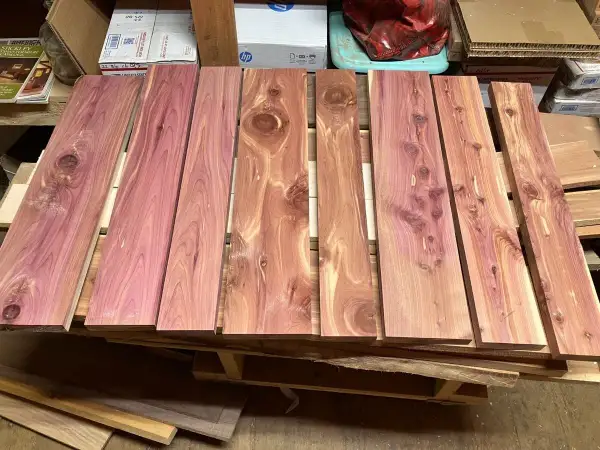 Tablones de madera de enebro con vetas rosadas y nudos visibles, apilados sobre un palé.